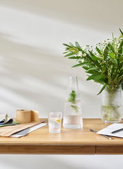 Pilastro Karaffe mit Glas, 1 l, auf einem Tisch mit Blumen und Schreibmaterialien, seitliche Perspektive.