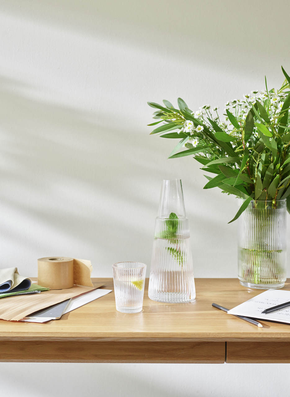 Pilastro Karaffe mit Glas, 1 l, auf einem Tisch mit Blumen und Schreibmaterialien, seitliche Perspektive.