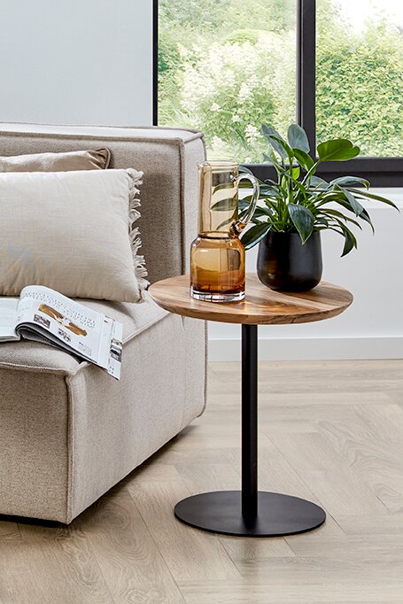 Moderner runder Beistelltisch mit Holzplatte und schwarzem Metallfuß neben beigem Sofa, darauf Glasvase und schwarze Pflanzschale, in seitlicher Perspektive auf Augenhöhe