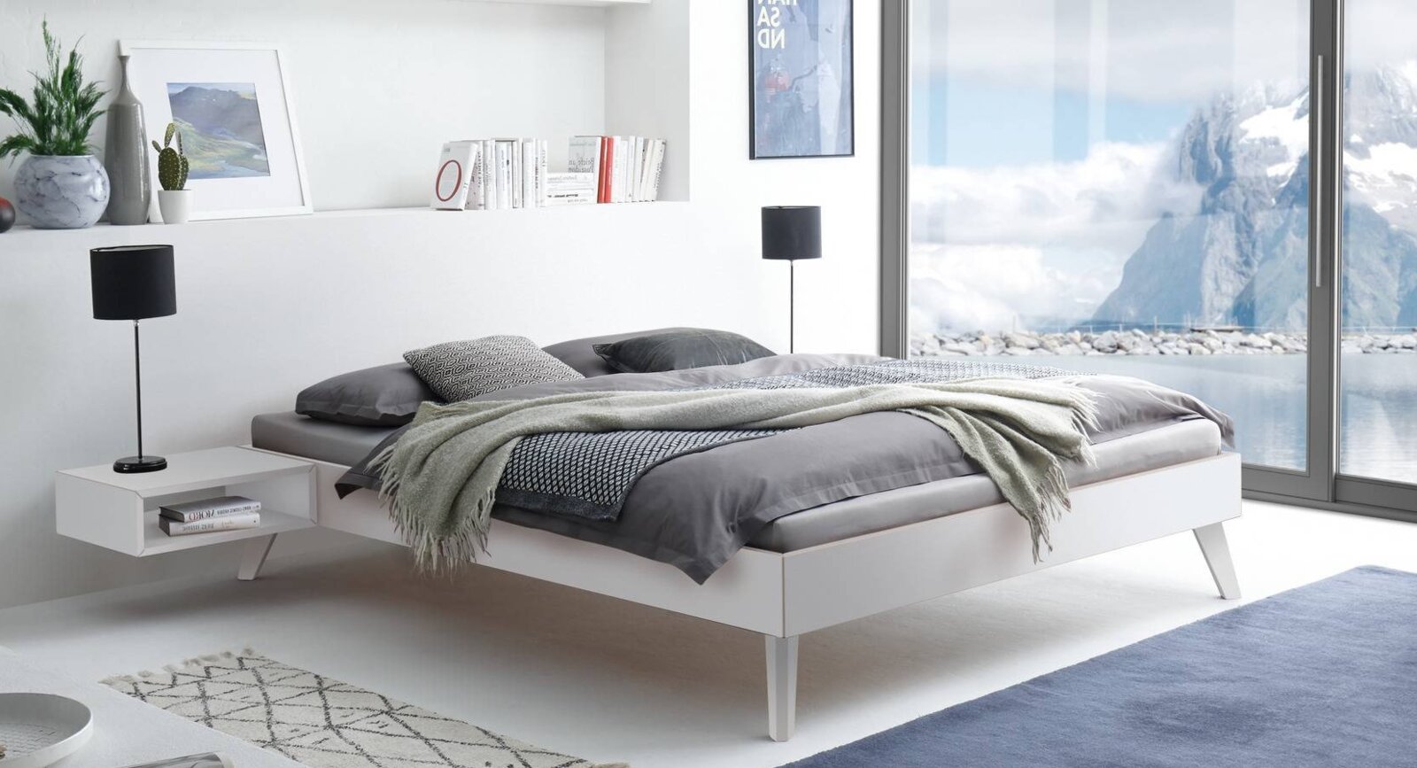 Modern wit bedframe met schuine poten en grijs beddengoed in een lichte slaapkamer, perspectivisch zijaanzicht vanaf rechtsvoor.