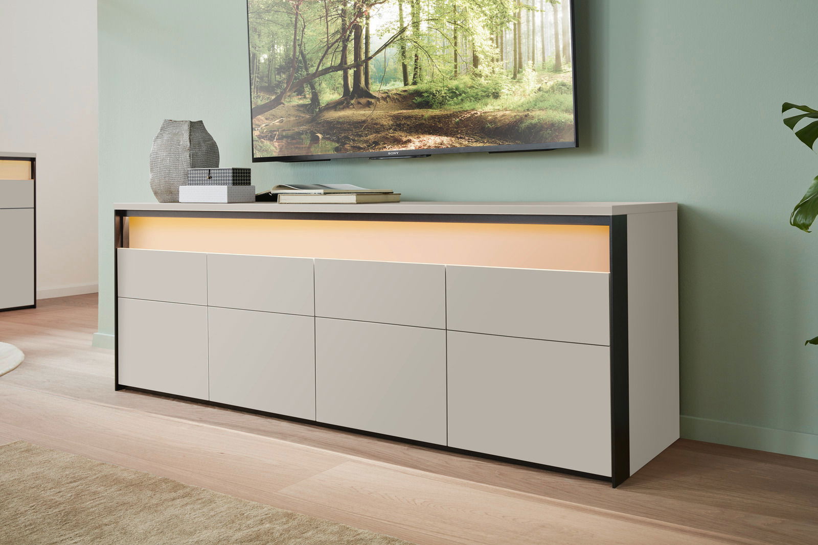 Modernes TV-Lowboard in hellem Holz mit schwarzem Rahmen, seitliche Perspektive, dekoriert mit Vasen und Büchern, unter einem an der Wand montierten Fernseher.