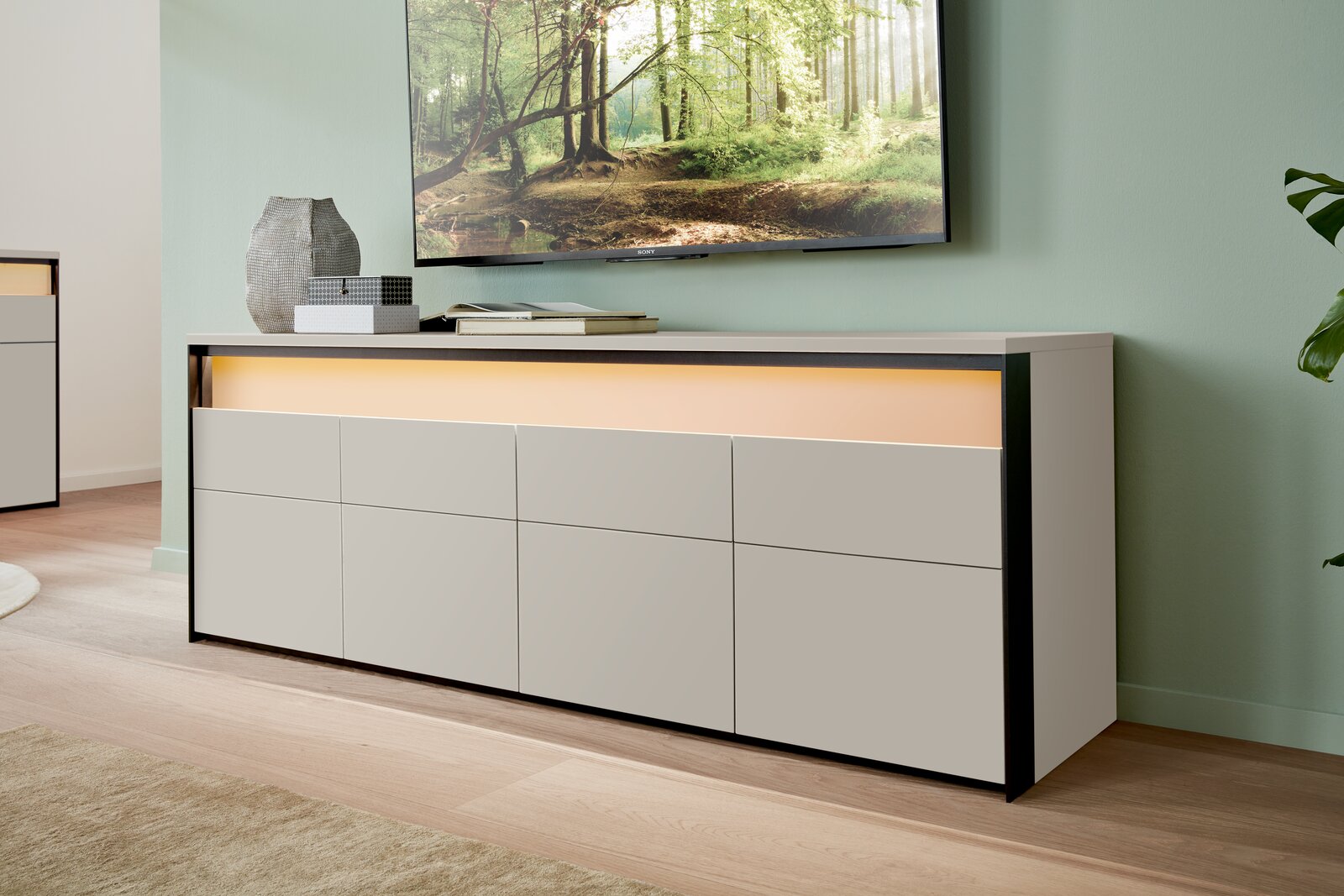 Modernes TV-Lowboard in hellem Holz mit schwarzem Rahmen, seitliche Perspektive, dekoriert mit Vasen und Büchern, unter einem an der Wand montierten Fernseher.