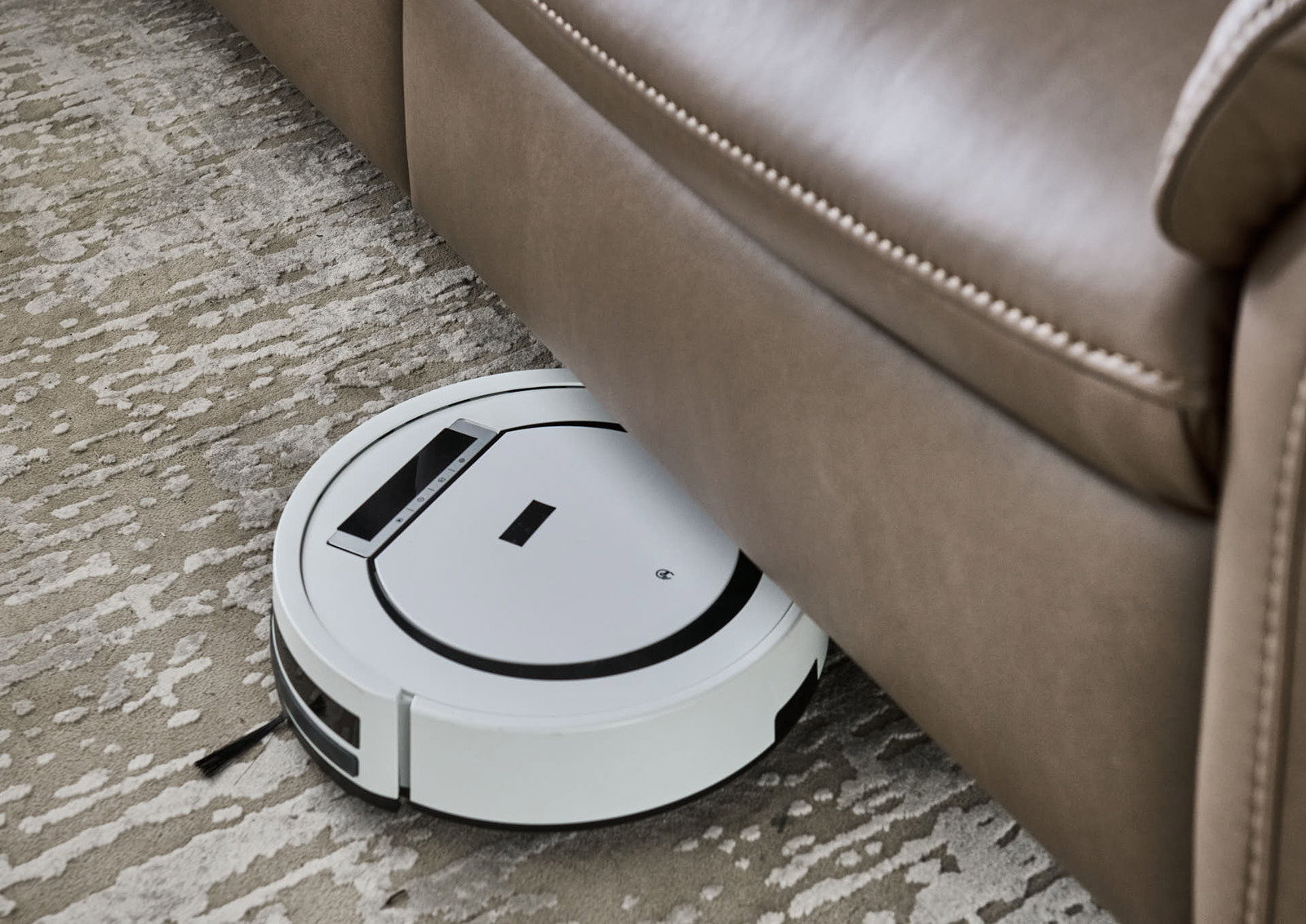 Ein weißer Saugroboter reinigt den Boden unter einem braunen Ledersofa aus der Vogelperspektive.
