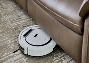 Een witte robotstofzuiger maakt de vloer schoon onder een bruine leren bank, gezien vanuit vogelperspectief.