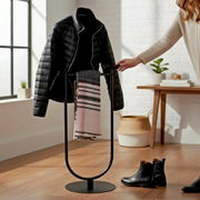 Schwarzer freistehender Kleiderständer mit runder Bodenplatte, darauf eine schwarze Steppjacke und ein rosa-grau gestreifter Schal; Innenraum-Szene aus frontaler Perspektive auf das Produkt.