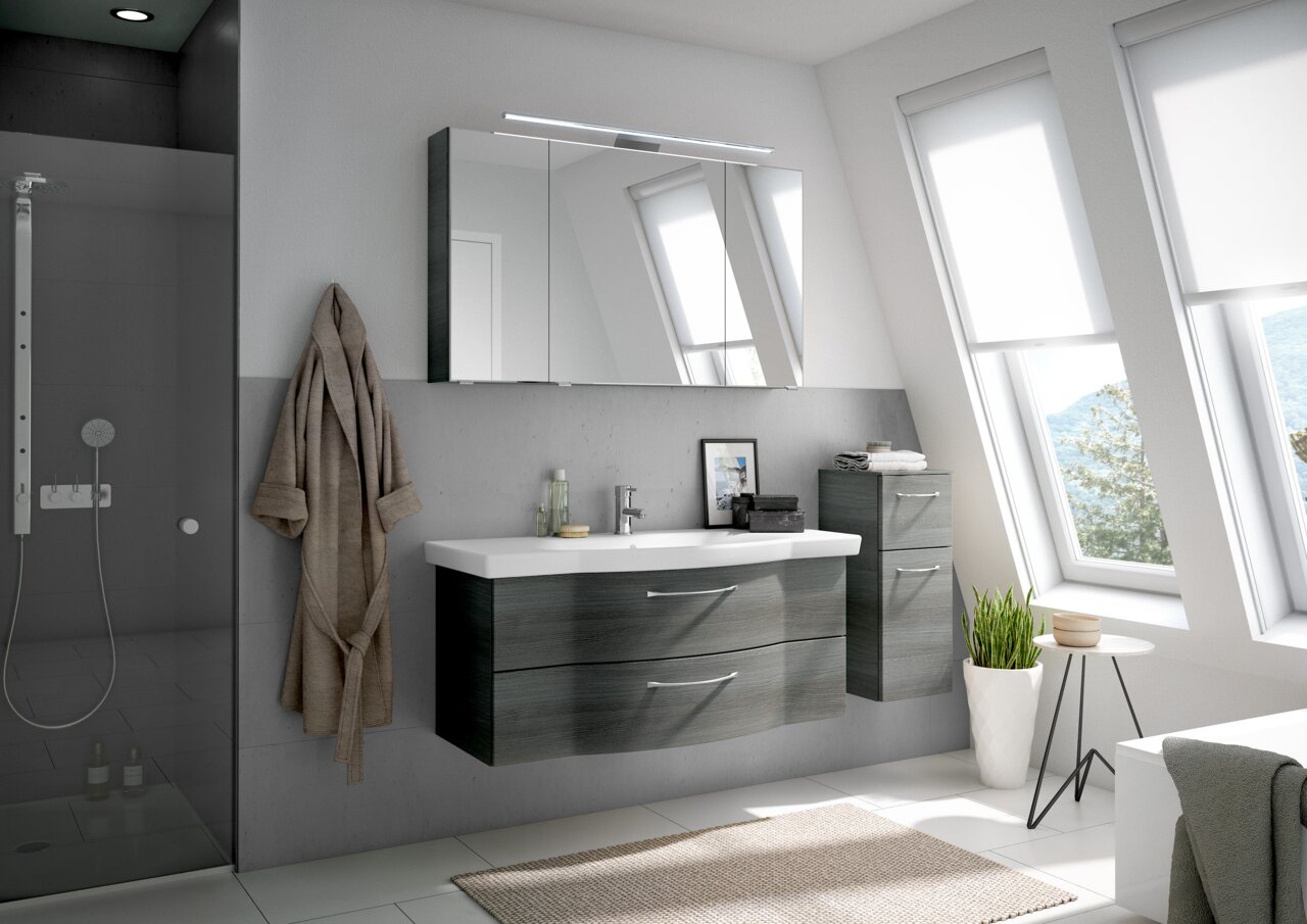 Moderne Badezimmermöbel in einem hellen Raum mit schrägen Fenstern, bestehend aus einem hängenden Waschtisch mit zwei Schubladen und einem Spiegelschrank darüber, seitlich ein hoher Schrank. Perspektive von vorne.