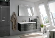 Moderne Badezimmermöbel in einem hellen Raum mit schrägen Fenstern, bestehend aus einem hängenden Waschtisch mit zwei Schubladen und einem Spiegelschrank darüber, seitlich ein hoher Schrank. Perspektive von vorne.