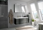 Moderne Badezimmermöbel in einem hellen Raum mit schrägen Fenstern, bestehend aus einem hängenden Waschtisch mit zwei Schubladen und einem Spiegelschrank darüber, seitlich ein hoher Schrank. Perspektive von vorne.