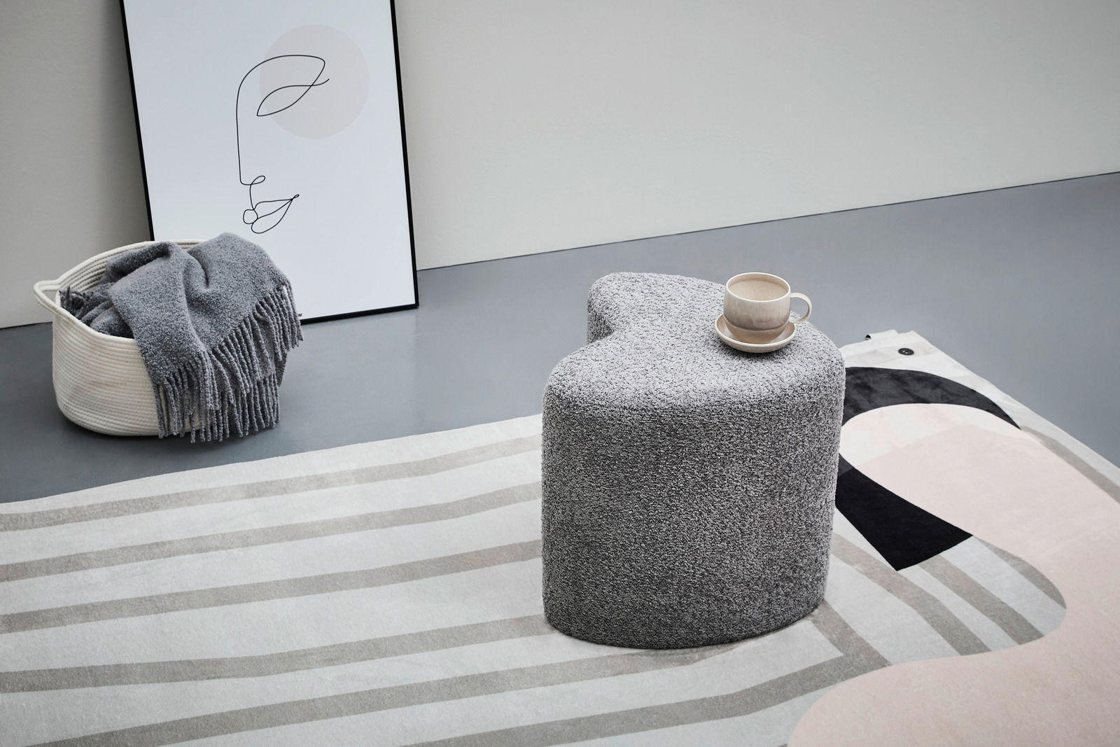 XORA Hocker ANIKA Grauer, herzförmiger Hocker XORA ANIKA in einer modernen Wohnumgebung, seitliche Perspektive, mit einer Tasse auf der Oberfläche.