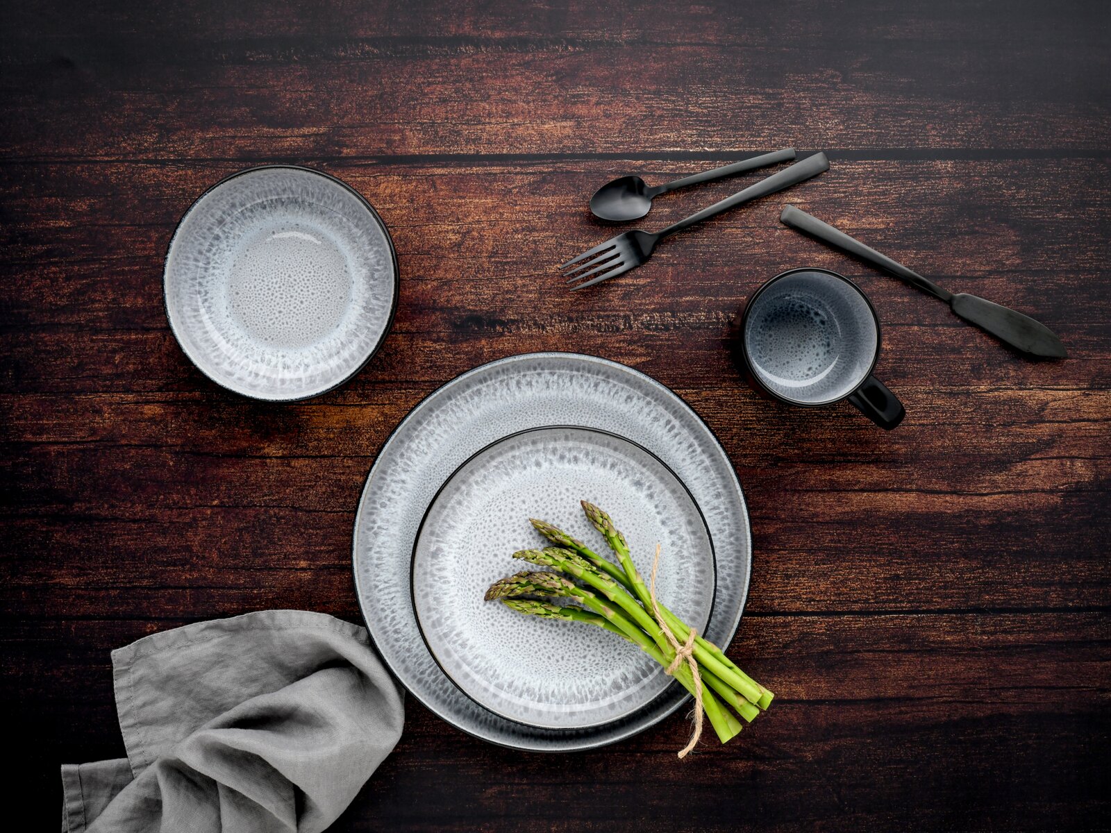 Draufsicht auf ein graues, strukturiertes Geschirr-Set mit zwei Tellern, Schale und Tasse auf dunkler Holzoberfläche, dazu schwarzes Besteck und ein Bund grüner Spargel.