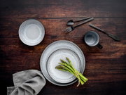 Draufsicht auf ein graues, strukturiertes Geschirr-Set mit zwei Tellern, Schale und Tasse auf dunkler Holzoberfläche, dazu schwarzes Besteck und ein Bund grüner Spargel.