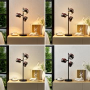 Moderne schwarze Tischlampe mit drei kugelförmigen, spiegelnden Schirmen in Roségold/Chrom auf Holz-Board, warm leuchtend neben Tasse, Büchern und Deko-Hirsch; frontale Perspektive auf dem Sideboard.