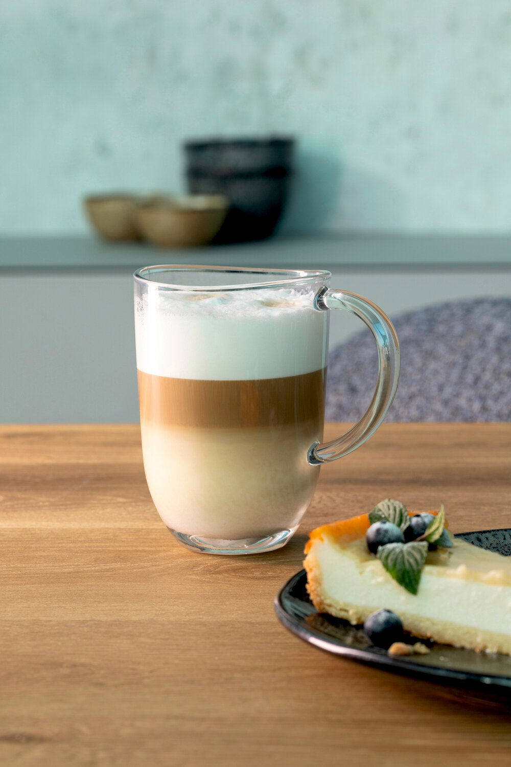 Leonardo Kaffeetasse NAPOLI Glas-Tasse Napoli 380ml mit Latte Macchiato, seitliche Perspektive, auf einem Holztisch neben einem Stück Käsekuchen.