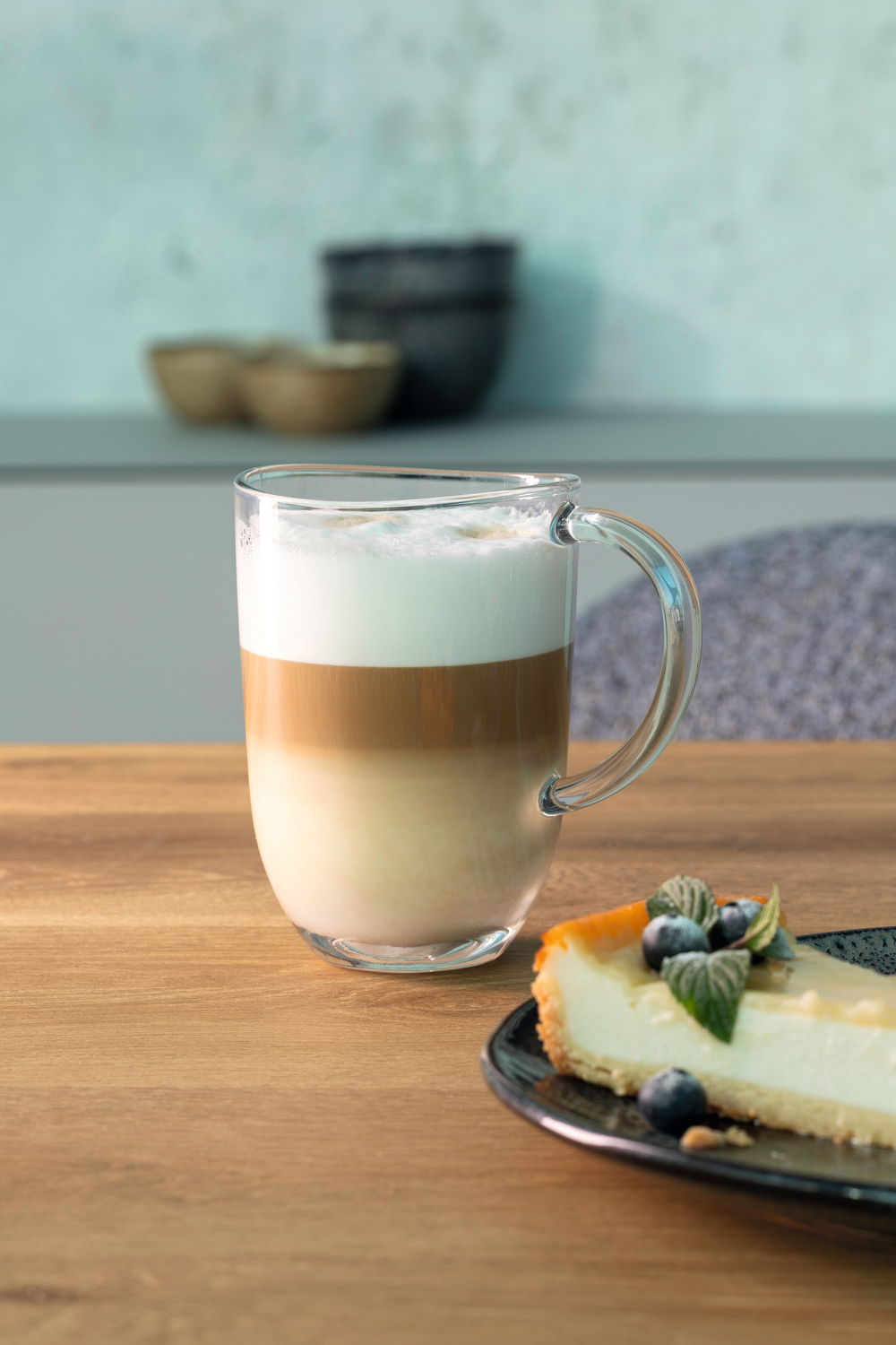 Glas-Tasse Napoli 380ml mit Latte Macchiato, seitliche Perspektive, auf einem Holztisch neben einem Stück Käsekuchen.