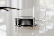 Stapel runder Metall-Untersetzer in Halterung auf glänzendem Tisch, mit Trinkglas im Hintergrund; Perspektive: leicht seitliche Nahaufnahme auf Augenhöhe.