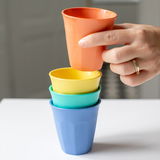 Vier bunte Becher in einem 300ml 4er-Set, gestapelt auf einem Tisch, mit einer Hand, die den obersten orangefarbenen Becher hält, aus der Seitenperspektive.