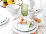 Schlichtes weißes Geschirr-Set mit welligem Rand (Teller, Schalen und Tassen) auf gedecktem Tisch mit grünen geriffelten Glasbecher und Macarons, aufgenommen in leicht erhöhter Frontansicht.