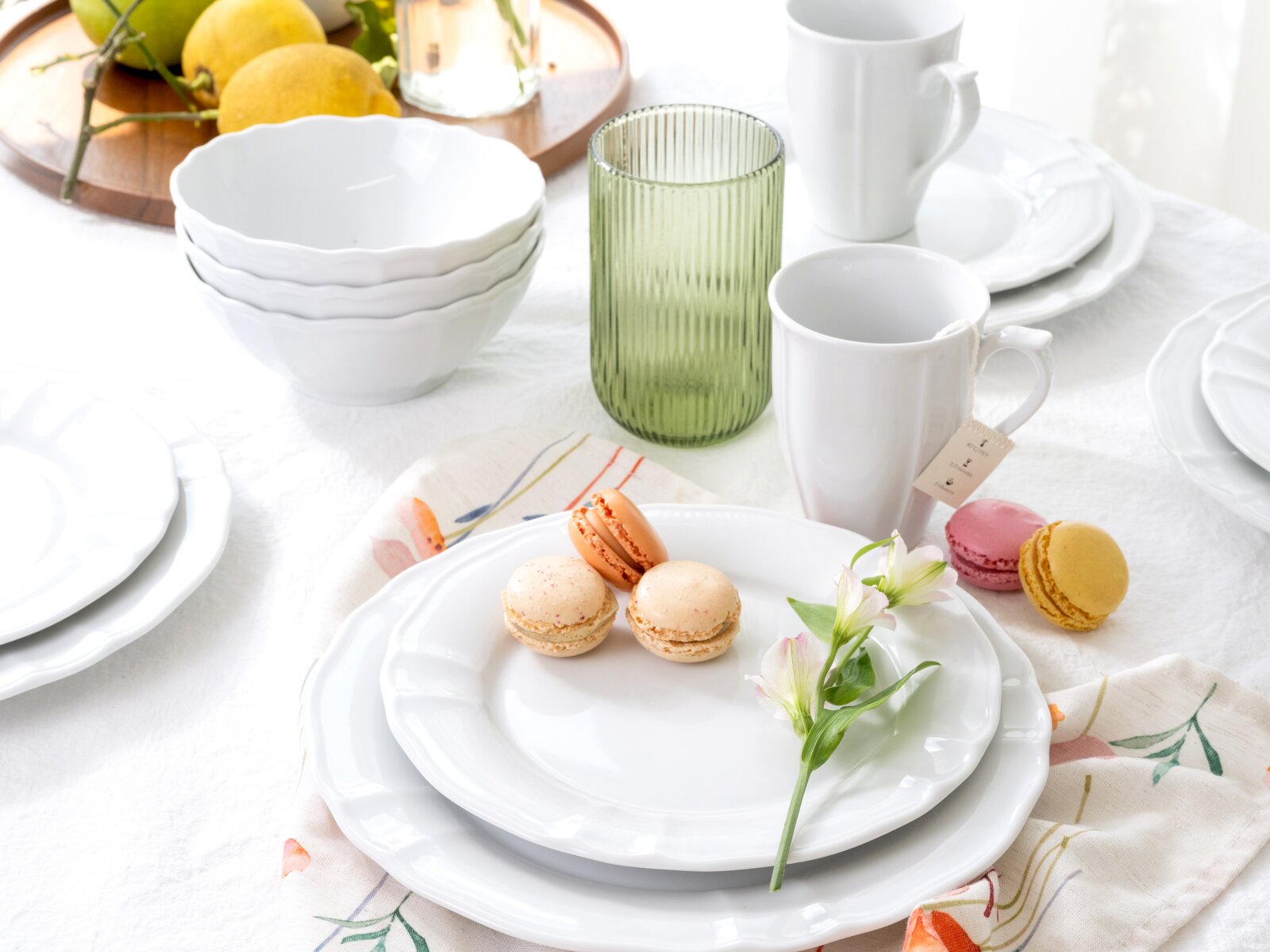Schlichtes weißes Geschirr-Set mit welligem Rand (Teller, Schalen und Tassen) auf gedecktem Tisch mit grünen geriffelten Glasbecher und Macarons, aufgenommen in leicht erhöhter Frontansicht.