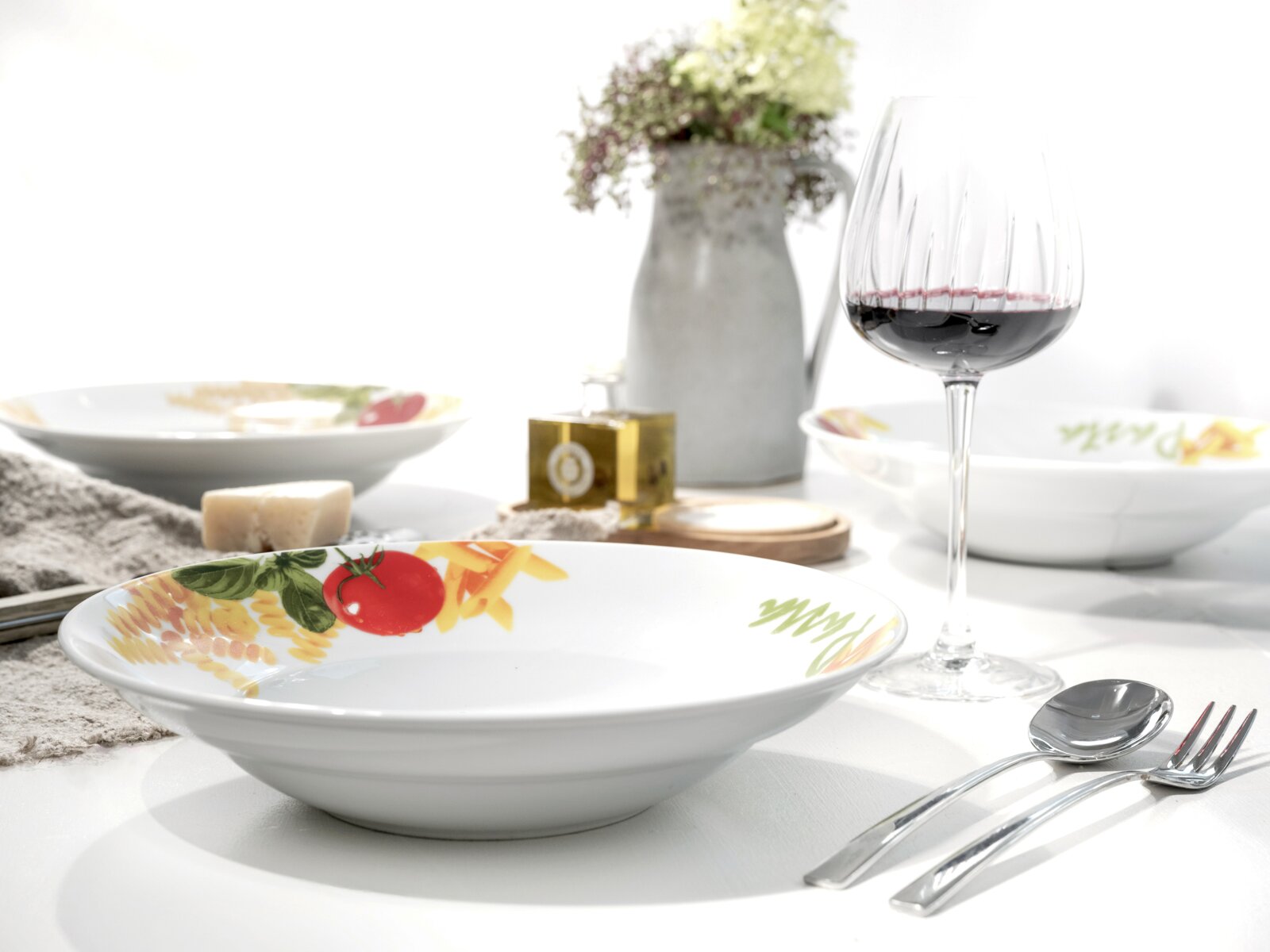 Weißer Suppenteller mit farbigem Gemüse- und Pasta-Dekor am Rand auf gedecktem Tisch, fotografiert aus leicht schräger Frontperspektive.