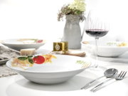 Weißer Suppenteller mit farbigem Gemüse- und Pasta-Dekor am Rand auf gedecktem Tisch, fotografiert aus leicht schräger Frontperspektive.