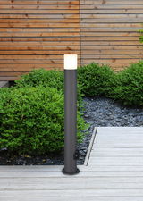 Moderne Außenleuchte in zylindrischer Form mit E27-Fassung, aufgestellt auf einer Terrasse mit Holzboden und umgeben von grünen Büschen, seitliche Perspektive.