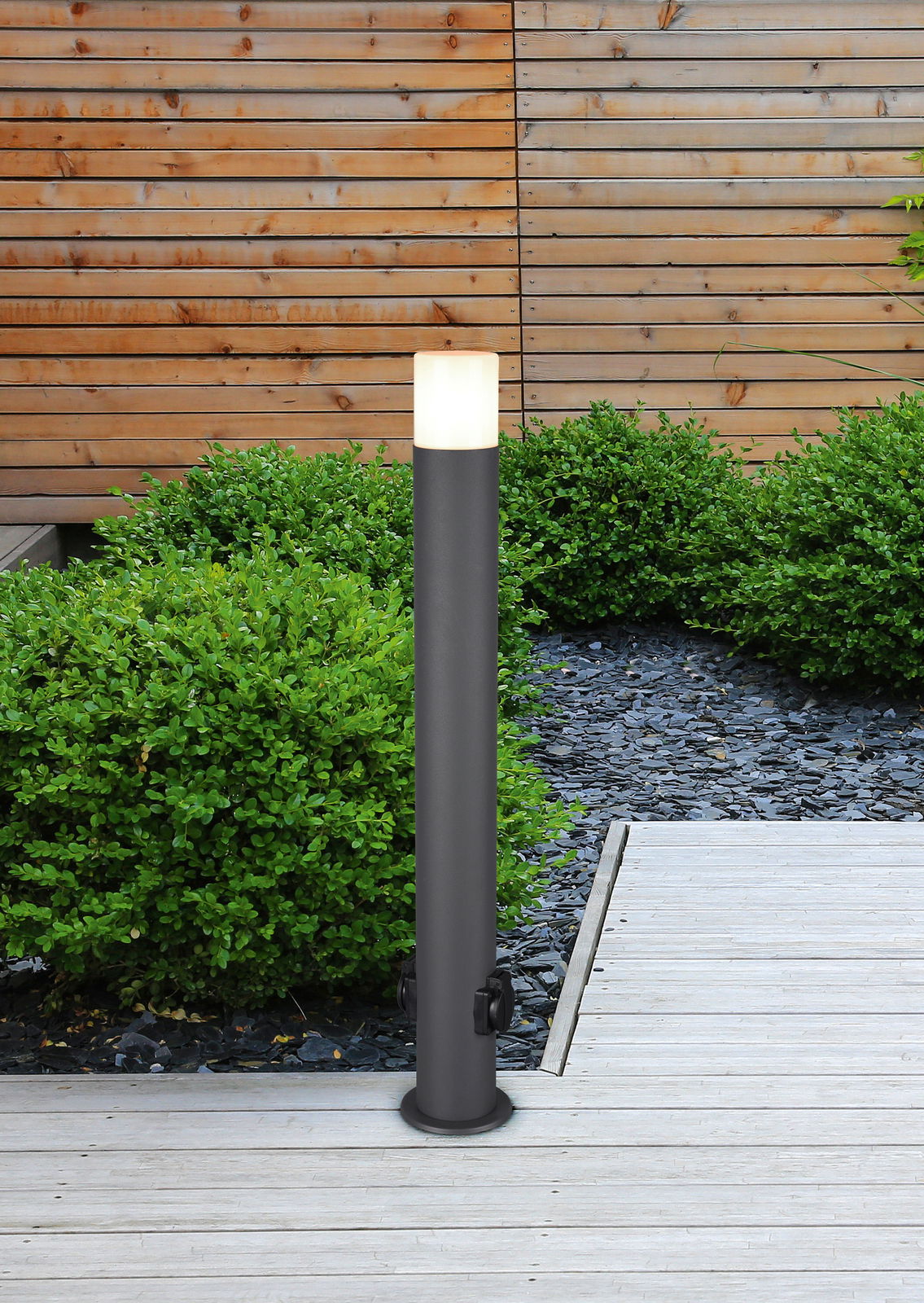 Moderne Außenleuchte in zylindrischer Form mit E27-Fassung, aufgestellt auf einer Terrasse mit Holzboden und umgeben von grünen Büschen, seitliche Perspektive.