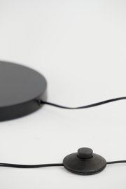 Close-up van de voet van een staande lamp met een zwarte voetschakelaar en kabel, gezien vanaf de zijkant.