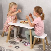 Zwei Kinder sitzen an einem kleinen weißen Kindertisch mit Holzbeinen und passender Bank und essen Snacks; seitliche Perspektive auf Tisch und Sitzbank.