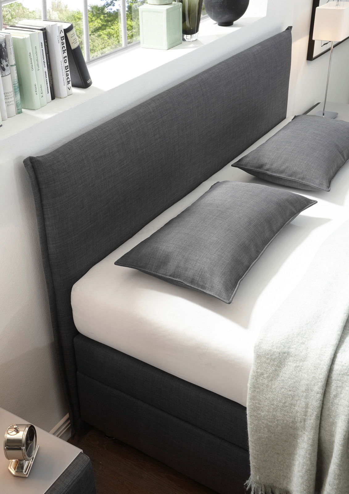Seitliche Perspektive eines stilvollen Boxspringbetts mit grauem Stoffbezug, zwei passenden Kissen und einer weißen Matratze, umgeben von moderner Dekoration.