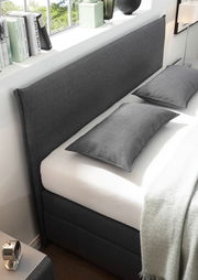 Seitliche Perspektive eines stilvollen Boxspringbetts mit grauem Stoffbezug, zwei passenden Kissen und einer weißen Matratze, umgeben von moderner Dekoration.