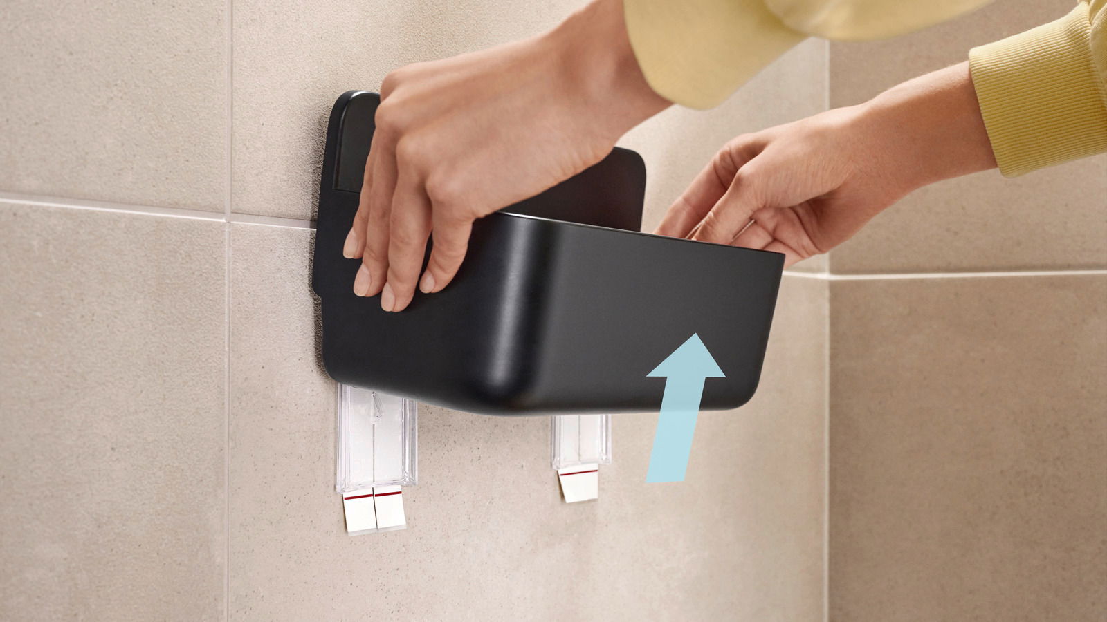Nahaufnahme eines schwarzen Duschkorbs von tesa Lavaa, der an einer gefliesten Wand montiert wird. Hände zeigen, wie der Korb in die Halterung eingesetzt wird.