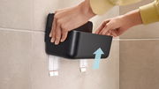 Nahaufnahme eines schwarzen Duschkorbs von tesa Lavaa, der an einer gefliesten Wand montiert wird. Hände zeigen, wie der Korb in die Halterung eingesetzt wird.