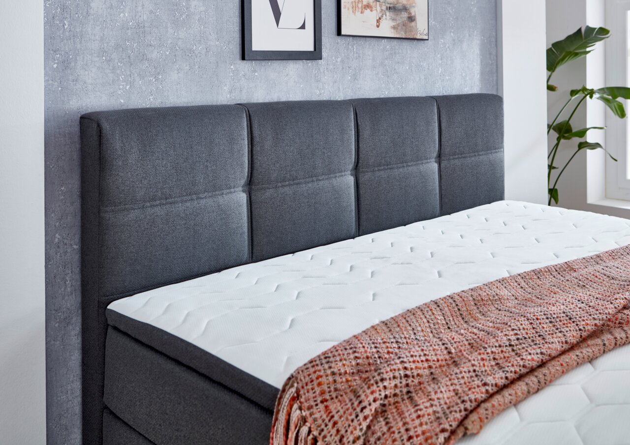 Boxspringbett mit grauem Kopfteil und weißer Matratze, seitliche Perspektive
