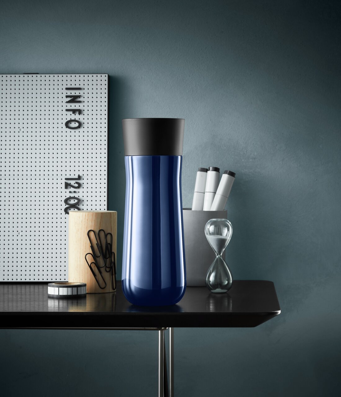 Blauer Impulse Isolierbecher 0,35l mit schwarzem Deckel, frontal auf einem Tisch mit Büroaccessoires.