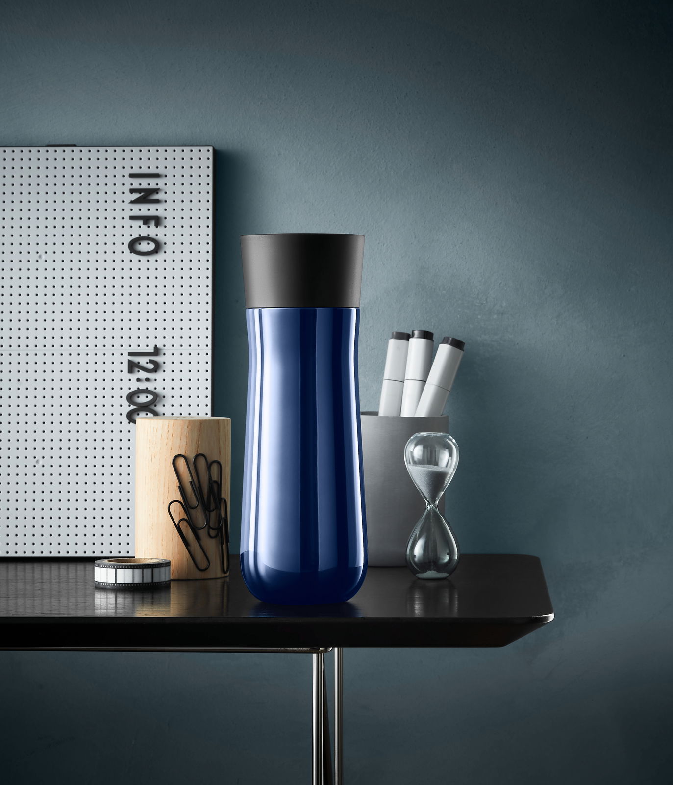 Blauer Impulse Isolierbecher 0,35l mit schwarzem Deckel, frontal auf einem Tisch mit Büroaccessoires.