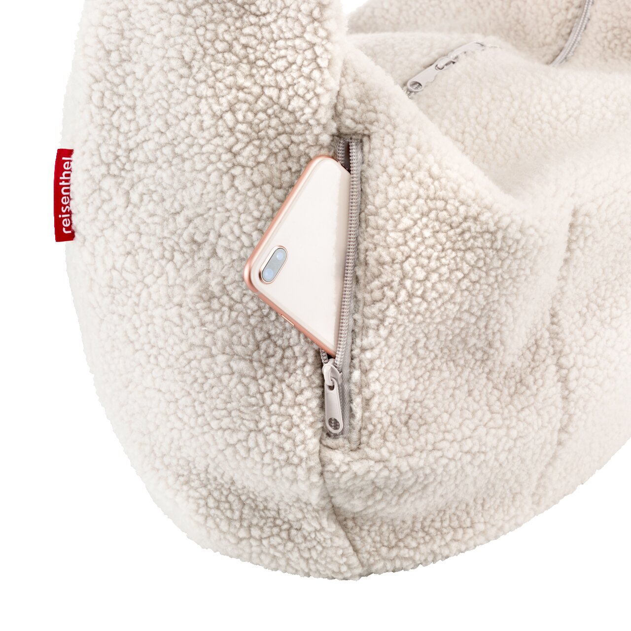 Close-up van een crèmekleurige tas van teddyfleece met een vak met ritssluiting, waaruit een smartphone steekt; perspectief: detailopname vanaf de zijkant.