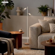 Weiße, schlanke Stehleuchte mit zwei verstellbaren Spot-Köpfen in modernem Wohnzimmer neben beigem Sofa, Frontansicht auf Augenhöhe