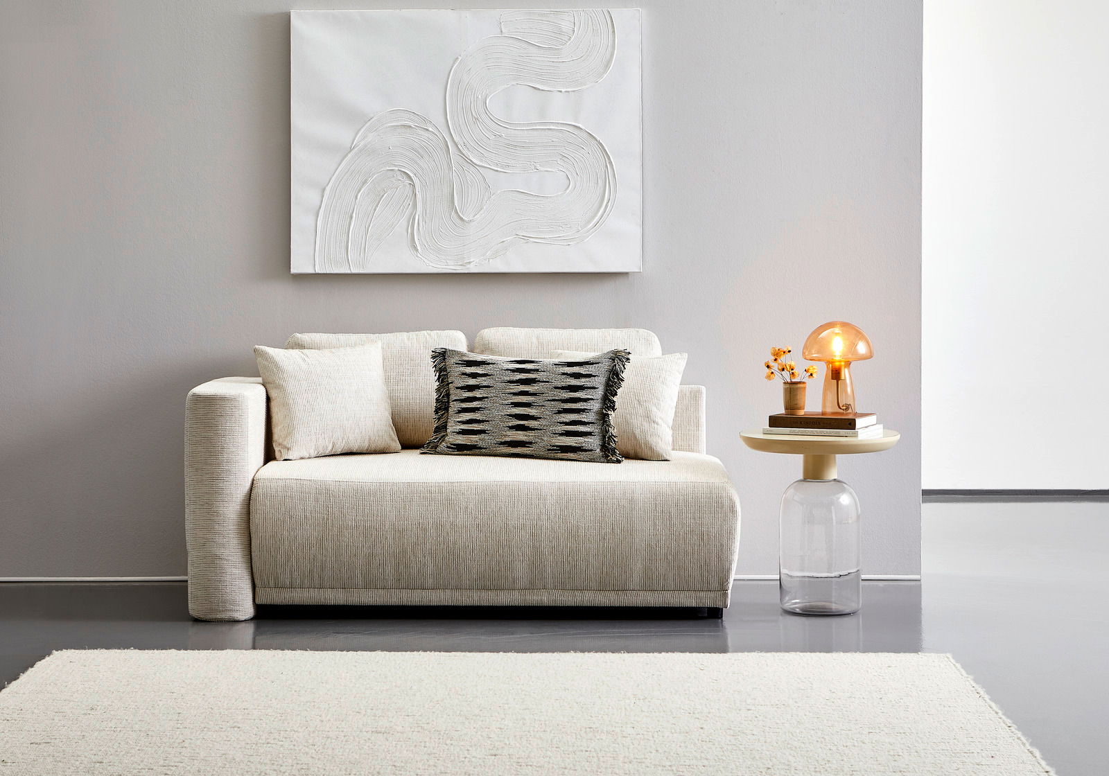 Beige Recamiere mit Kissen, frontal fotografiert, neben einem runden Beistelltisch mit Lampe und Dekoration.