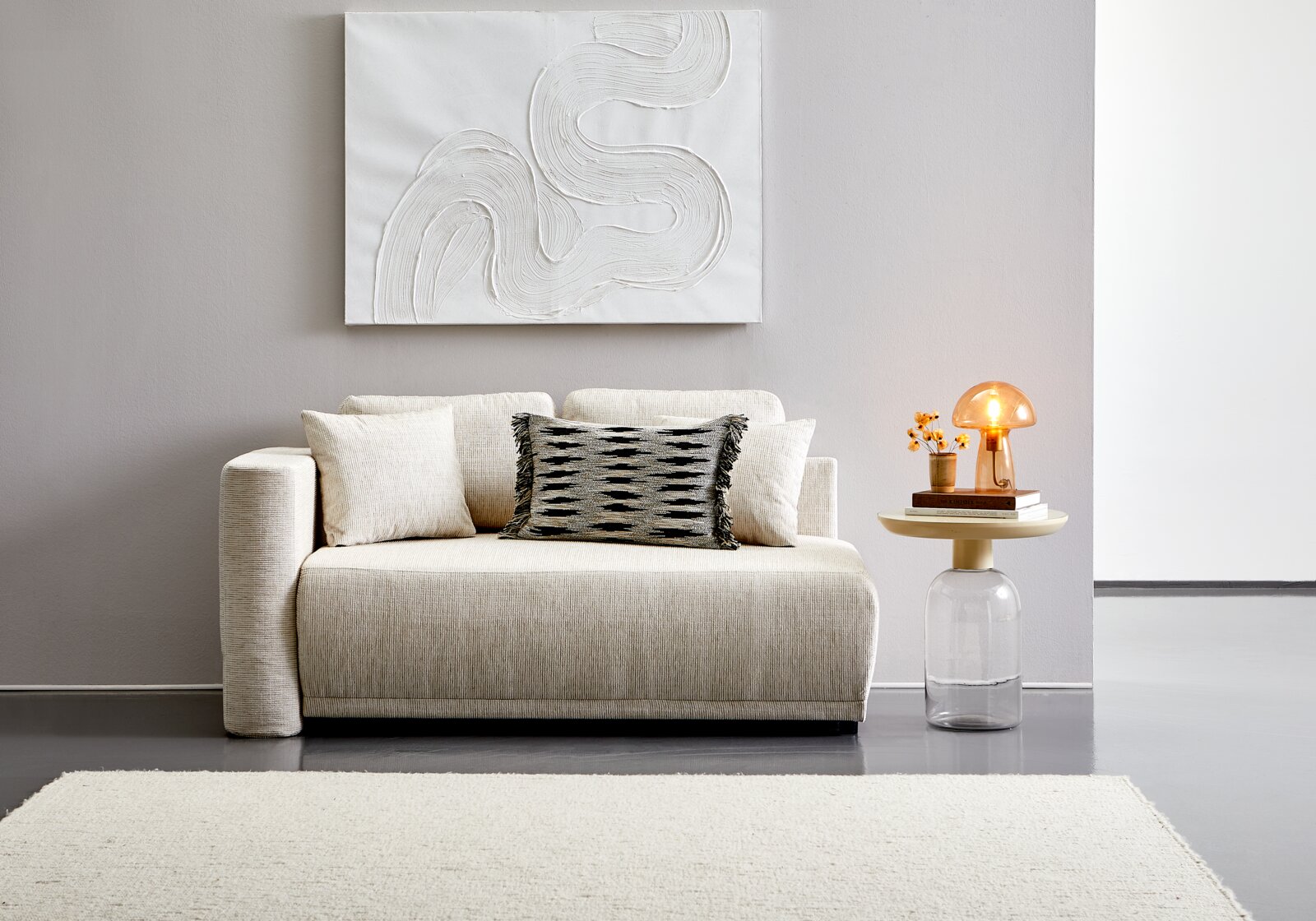 Beige Recamiere mit Kissen, frontal fotografiert, neben einem runden Beistelltisch mit Lampe und Dekoration.
