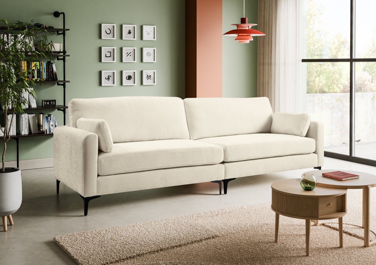 Beiges 3-Sitzer-Sofa mit Stoffbezug und schwarzen Metallfüßen im modernen Wohnzimmer, frontale leicht schräge Perspektive von links im Hintergrund grüne Wand mit Bildern, Regal und rote Pendelleuchte, rechts Fenster und Couchtisch.