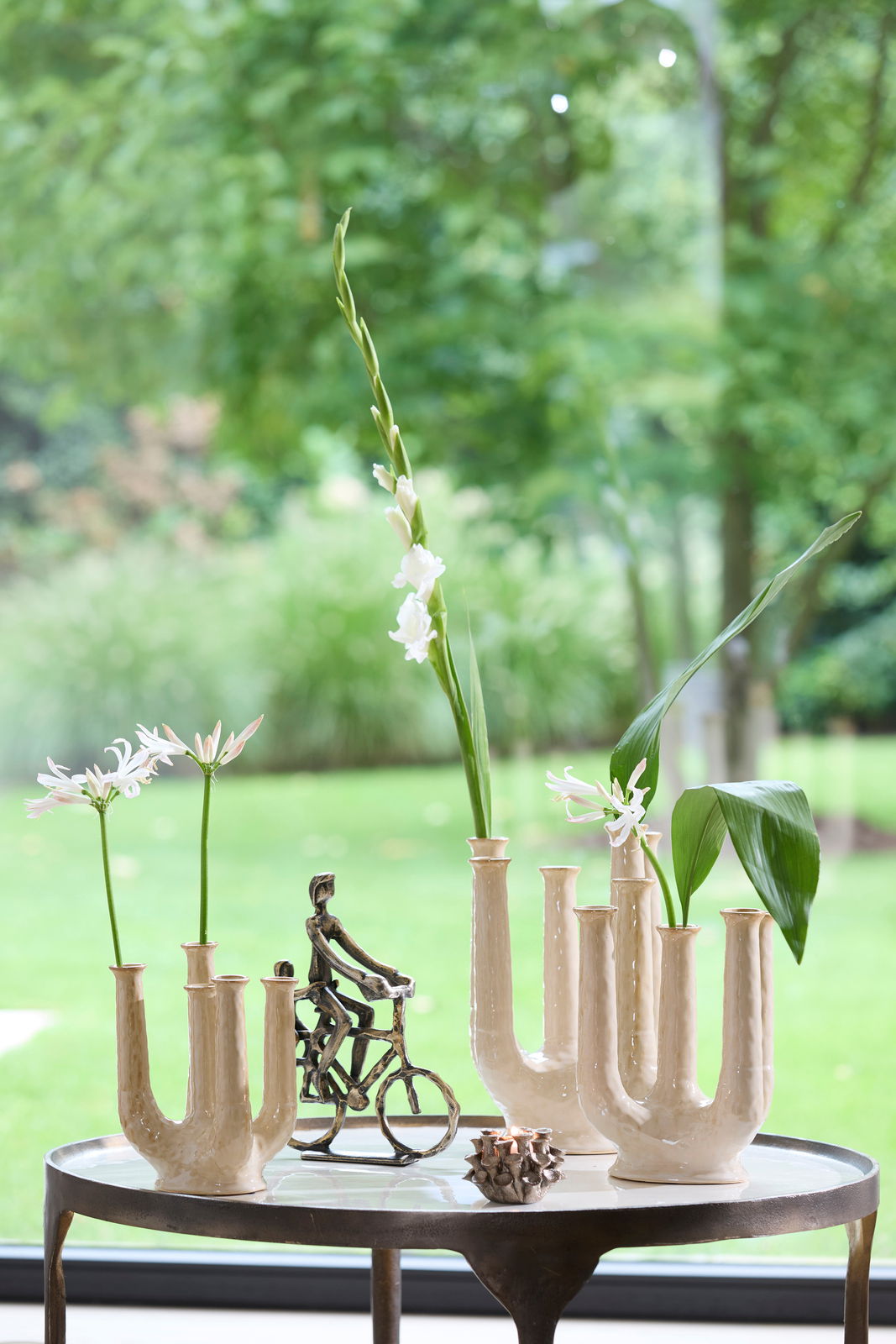 Ein stilvoller Teelichthalter auf einem Tisch, umgeben von dekorativen Vasen mit Blumen und einer kleinen Skulptur eines Radfahrers, mit Blick auf einen grünen Garten im Hintergrund.