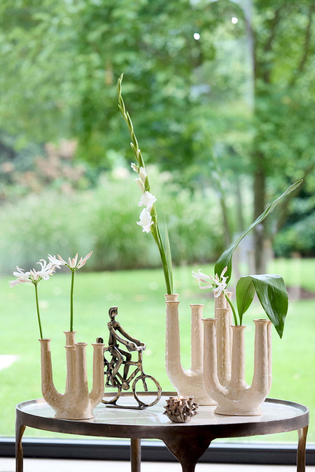 Ein stilvoller Teelichthalter auf einem Tisch, umgeben von dekorativen Vasen mit Blumen und einer kleinen Skulptur eines Radfahrers, mit Blick auf einen grünen Garten im Hintergrund.