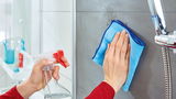 Eine Person reinigt eine Duschwand mit einem blauen Tuch und einem Sprühreiniger. Perspektive: Nahaufnahme der Reinigungshandlung.
