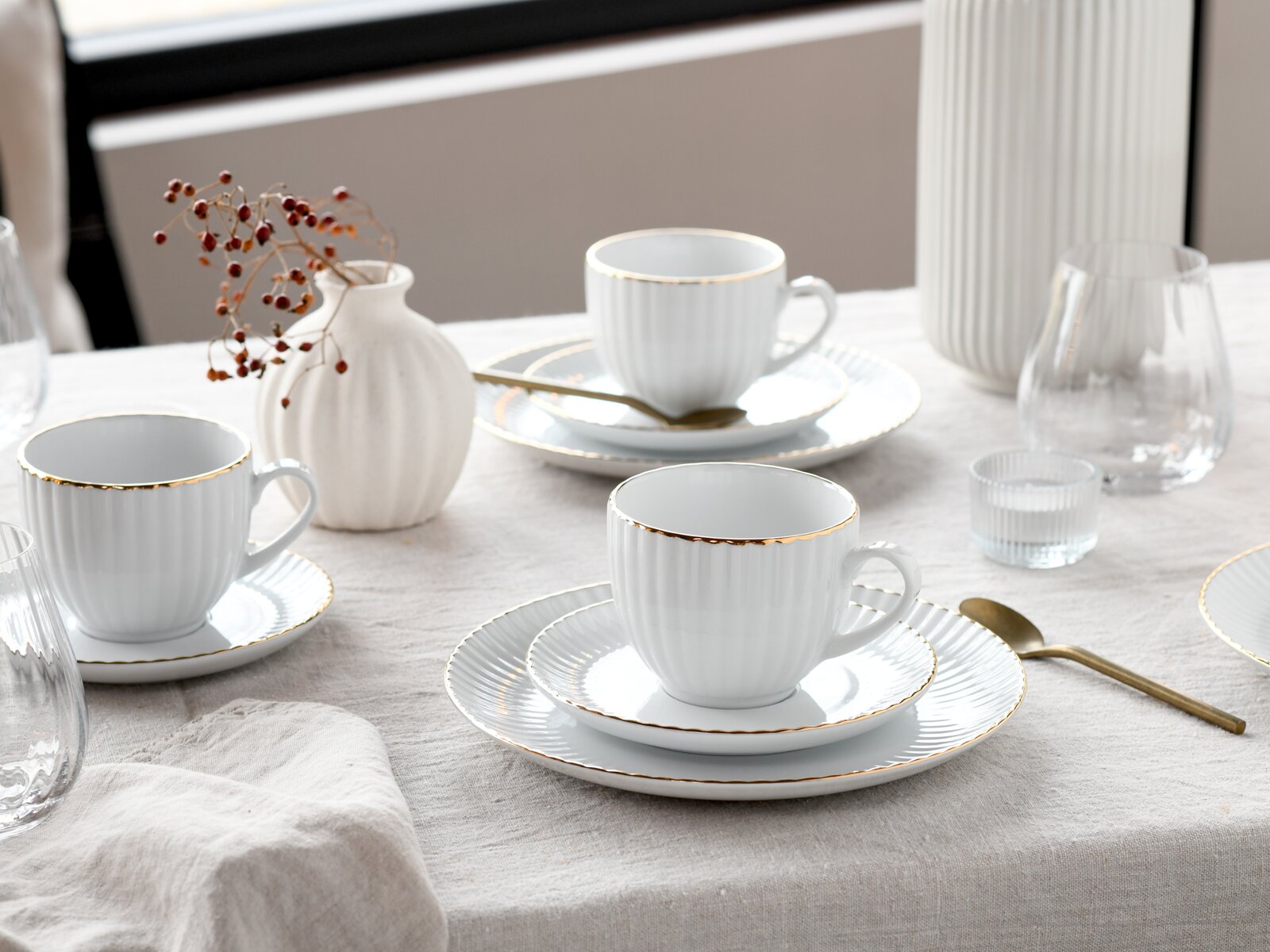 Schlichtes weißes Porzellan-Kaffeegeschirr mit feiner Goldkante (Tassen auf Untertassen und Tellern) auf hell gedecktem Tisch, fotografiert aus leicht erhöhter schräger Perspektive.