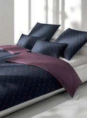 Bettwäsche-Set in dunklem Design mit geometrischem Muster, bestehend aus Kissen- und Bettbezug, auf einem Bett in einem hellen Raum, seitliche Perspektive.