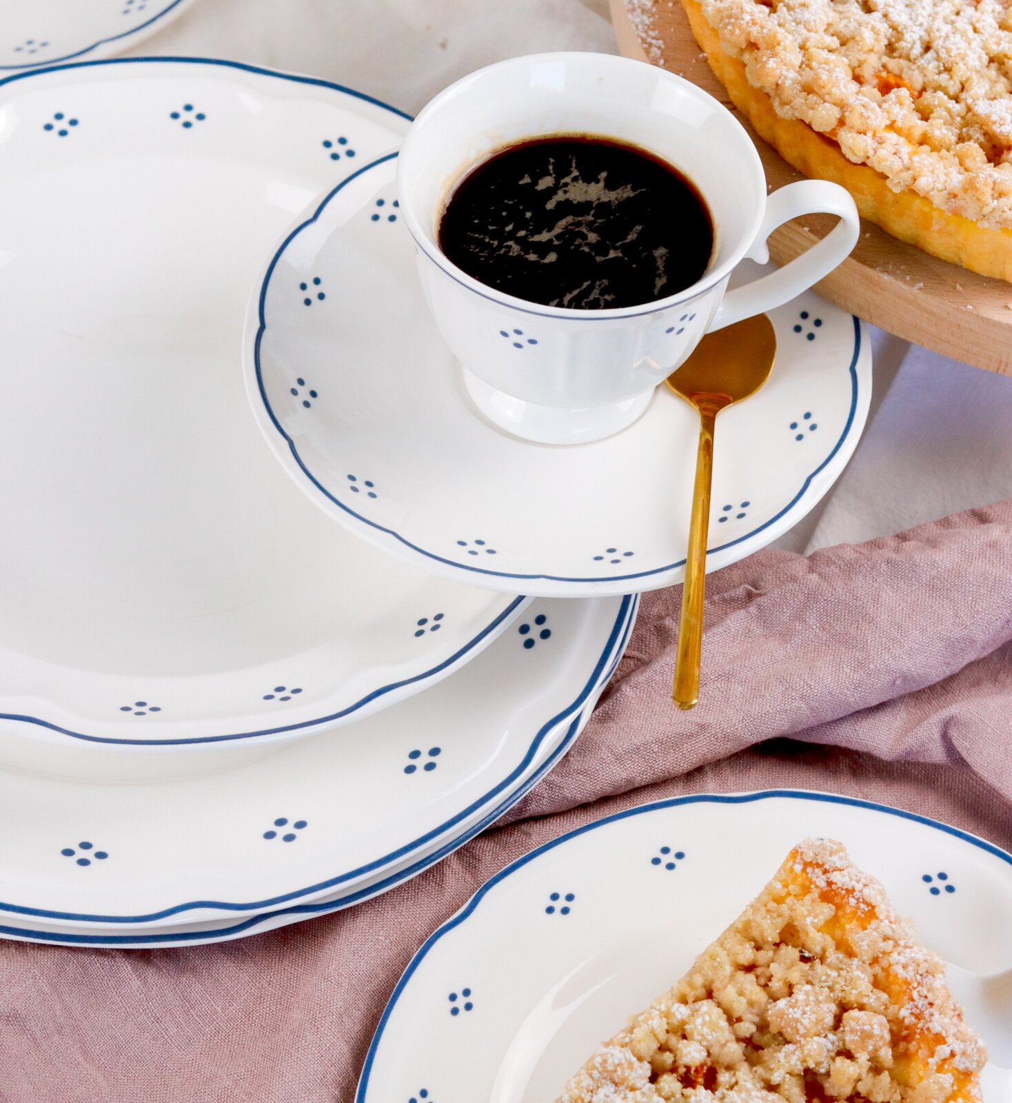 Schräg von oben fotografiertes, weißes Porzellan-Geschirrset mit blauen Punkten und Wellenrand Kaffeetasse mit schwarzem Kaffee auf Untertasse, goldener Löffel und Streuselkuchen im Hintergrund.
