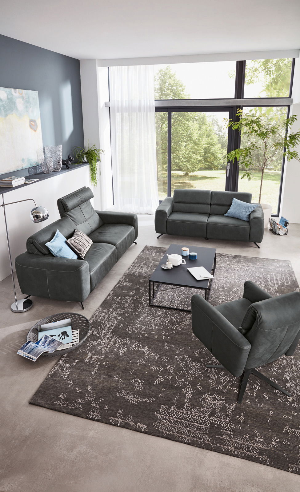 Modernes Wohnzimmer mit zwei dunkelgrauen Sofas und einem Sessel, von oben betrachtet. Große Fenster im Hintergrund lassen viel Licht herein.