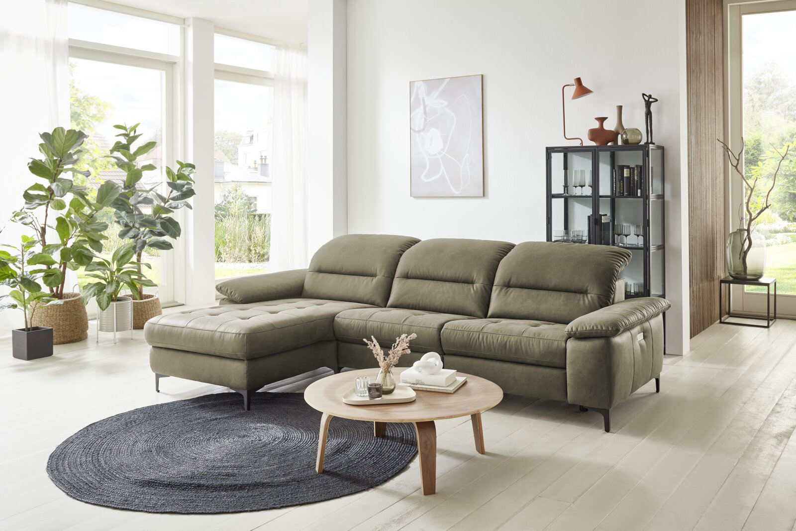 Grüne Longchairkombination in einem modernen Wohnzimmer mit großen Fenstern, Pflanzen und einem runden Holztisch. Perspektive von vorne links.
