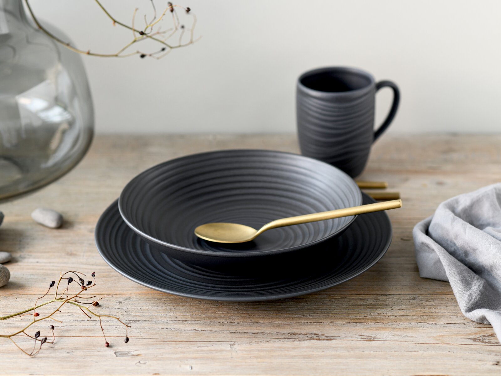 Creatable Kaffeebecher 6-tlg LAVA STONE Mattschwarze, geriffelte Keramikschale auf passendem Teller mit goldfarbenem Löffel, dahinter eine schwarze Tasse auf Holztisch; Perspektive: leicht von vorne, seitlich und von oben (3/4-Ansicht).