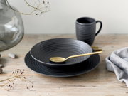 Mattschwarze, geriffelte Keramikschale auf passendem Teller mit goldfarbenem Löffel, dahinter eine schwarze Tasse auf Holztisch; Perspektive: leicht von vorne, seitlich und von oben (3/4-Ansicht).
